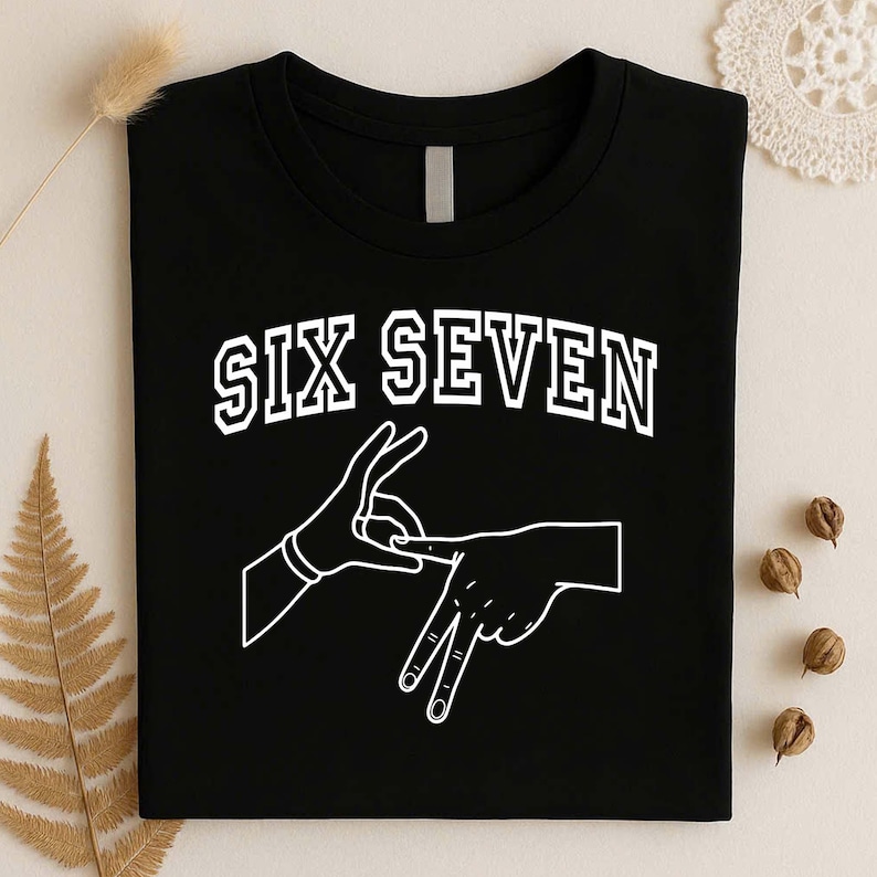 May include: A black t-shirt featuring the white text SIX SEVEN in a collegiate font arched above a white line-art graphic of two hands forming a hand sign. The design is centered on the chest of the shirt, which has a standard crew neckline.