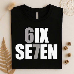 May include: A black t-shirt featuring the text 6IX SE7EN printed in a bold, sans-serif font. The number 6 is colored in light gray, while the remaining letters are white. The number 7 is also colored in light gray, creating a visual contrast.