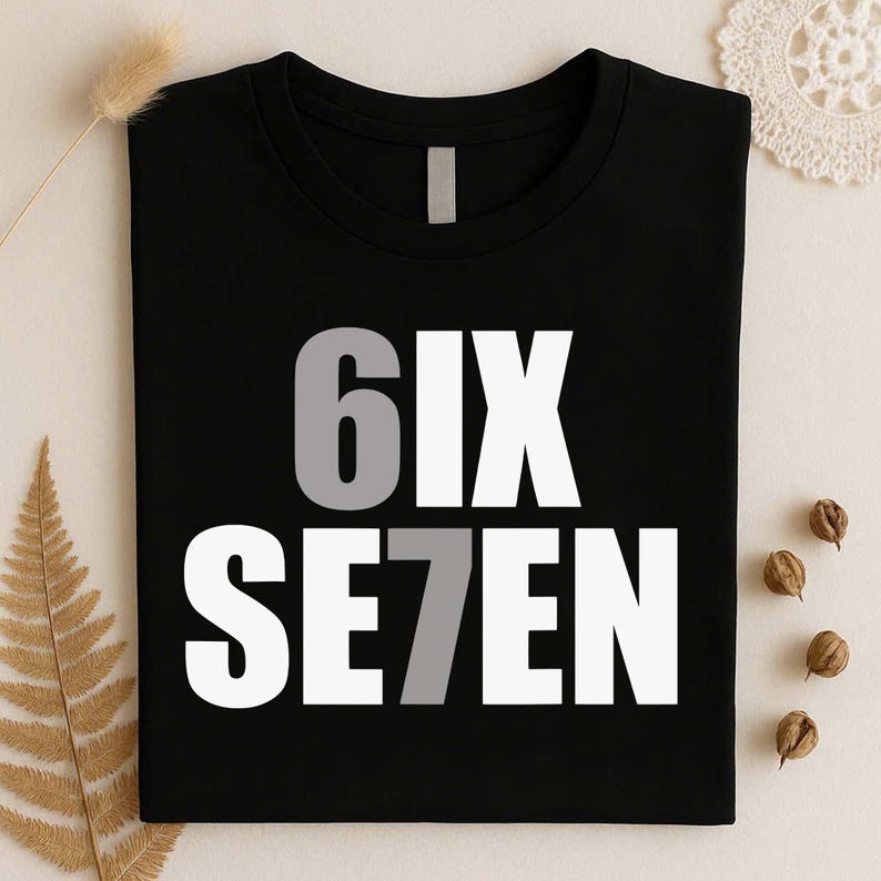 May include: A black t-shirt featuring the text 6IX SE7EN printed in a bold, sans-serif font. The number 6 is colored in light gray, while the remaining letters are white. The number 7 is also colored in light gray, creating a visual contrast.