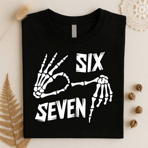 May include: A black t-shirt featuring a white graphic of two skeleton hands forming a hand gesture. The text "SIX" is printed above the hands, and the text "SEVEN" is printed below the hands in a bold, stylized white font on the front of the shirt.