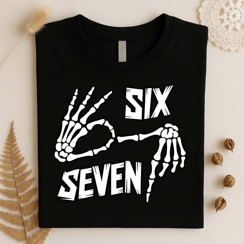 May include: A black t-shirt featuring a white graphic of two skeleton hands forming a hand gesture. The text "SIX" is printed above the hands, and the text "SEVEN" is printed below the hands in a bold, stylized white font on the front of the shirt.