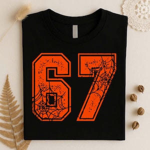 May include: A black crew neck t-shirt featuring a large, bold orange number 67 graphic on the front. The number 67 is decorated with a black spiderweb pattern and a distressed, textured finish. The shirt has a simple, short-sleeved design.
