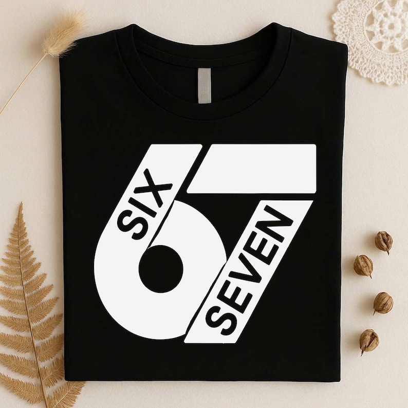 May include: A black short-sleeved t-shirt featuring a large white graphic design on the front. The graphic consists of the number 67 stylized with the words SIX and SEVEN integrated into the bold white shapes of the digits in black lettering.