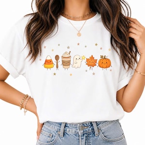 Op de afbeelding: Een wit t-shirt met korte mouwen en een grafische print van een candy corn, een houten lepel, een koffiebeker, een spookje met een ster, een herfstblad en een pompoen, allemaal met lachende gezichtjes, omringd door kleine gouden sterretjes.