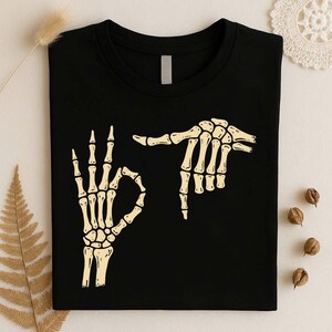 May include: A black crew neck t-shirt featuring a graphic print of two skeletal hands in a light cream color. One hand is positioned in an okay gesture, while the other hand is positioned horizontally above it, pointing toward the first hand.