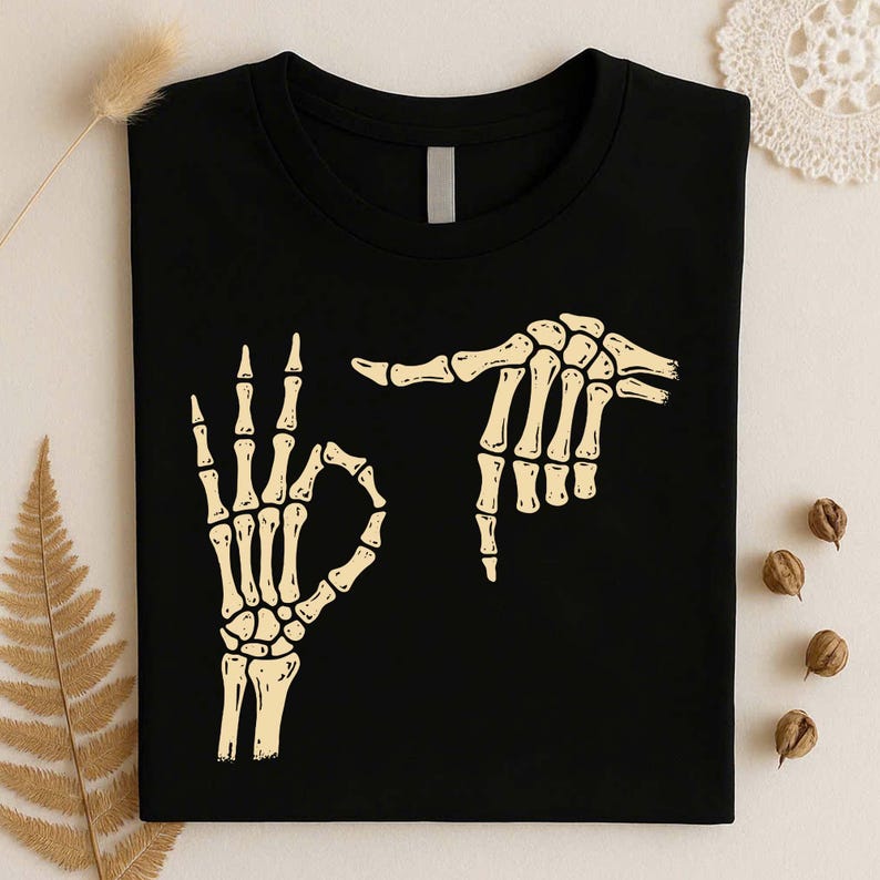 May include: A black crew neck t-shirt featuring a graphic print of two skeletal hands in a light cream color. One hand is positioned in an okay gesture, while the other hand is positioned horizontally above it, pointing toward the first hand.