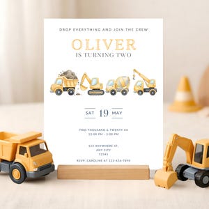 May include: A birthday invitation with a construction theme. The invitation features illustrations of construction vehicles and the text "OLIVER IS TURNING TWO". The invitation is propped up by a wooden stand, with toy construction vehicles in the foreground.