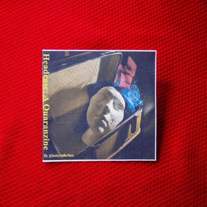 May include: A square sticker with the text "Headcase: A Quaranzine" and a small illustration of a plaster head inside a suitcase. The head has blue hair. The sticker is on a red textured surface.