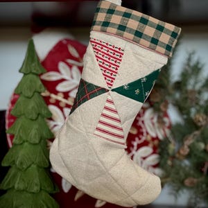 以下が含まれることがあります： ベージュの背景とパッチワークデザインのキルティングクリスマスストッキング。パッチワークには、赤と白のストライプ、緑の格子縞、緑と白の花柄が含まれています。ストッキングの上部には、緑とタンの格子縞模様があります。