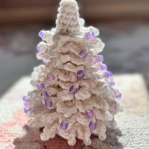 May include: A handmade, off-white crocheted Christmas tree adorned with small, translucent purple beads. The tree has a textured, layered appearance, with a conical shape and a slightly blurred background.