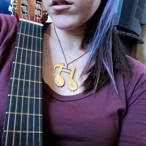 May include: A woman with purple hair wearing a maroon shirt and a gold musical note necklace. She is holding a brown acoustic guitar with a light wood fretboard.