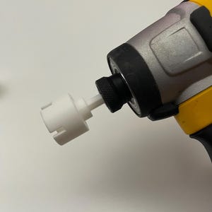 May include: A close-up of a yellow, black, and silver power drill with a white attachment. The drill is angled towards the viewer, showcasing its design and the attached tool. The background is a plain white surface.