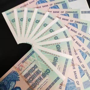 Op de afbeelding: Een waaier van Zimbabwaanse biljetten van 100 biljoen dollar. De lichtgroene biljetten tonen de tekst "RESERVE BANK OF ZIMBABWE" en "ONE HUNDRED TRILLION DOLLARS". De bankbiljetten zijn in een halve cirkel gerangschikt tegen een donkere achtergrond.