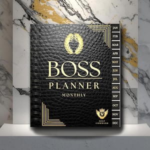 May include: A black leather-textured "BOSS PLANNER MONTHLY" notebook with gold lettering and accents. The cover features a gold laurel wreath and a crown emblem. The planner includes a monthly calendar and is displayed on a white surface.