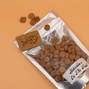 May include: A silver pouch filled with brown, round pet food, with a rectangular biscuit featuring a fishbone design. The words "Adventure On The Trail" are on a white label. Several pieces of food are scattered around.