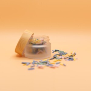 May include: A small, frosted glass jar with a wooden lid, partially open, revealing colorful sprinkles. The sprinkles are scattered around the jar on a light orange background. The jar is likely used for storing small items.