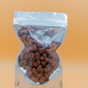 May include: A clear, silver-lined plastic bag filled with heart-shaped, brown treats. The bag is sealed at the top and has a curved shape, with a light orange background.