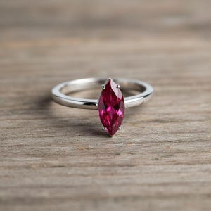 Puede incluir: Un anillo de plata con una gema rosa intenso con corte marquesa. La gema es el punto focal, engastada en una banda simple y elegante. El anillo se exhibe sobre una superficie de madera.