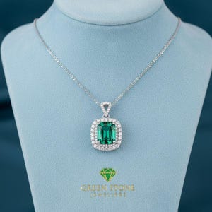 May include: A silver necklace with a square, emerald-cut green gemstone pendant, surrounded by a halo of smaller white gemstones. The pendant hangs from a delicate chain. The necklace is displayed on a blue surface. The text "GREEN STONE JEWELLERS" is at the bottom.