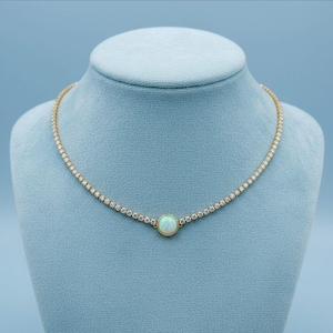 May include: A gold-toned necklace with a delicate chain of small, clear stones. The necklace features a central, round opal stone. The jewelry is displayed on a blue mannequin, showcasing the necklace's design and sparkle.
