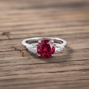 May include: A silver ring with a large, round, deep red gemstone as the focal point, flanked by two smaller, pear-shaped, clear stones. The ring is set against a wooden surface.