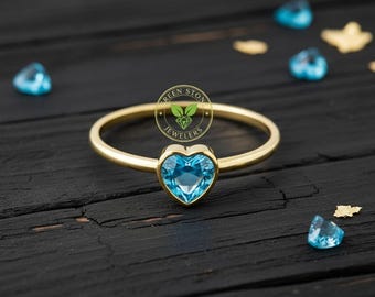 May include: A gold ring with a heart-shaped, light blue gemstone. The ring is set against a dark wooden surface, with scattered gemstones and gold flakes. The ring is a piece of jewelry.