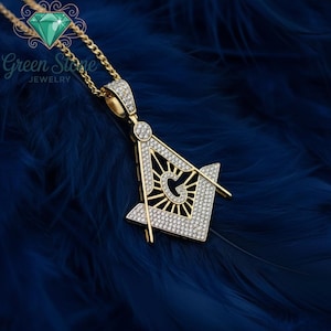 May include: Gold pendant necklace with a Masonic symbol, set with small, sparkling stones. The pendant hangs from a fine chain. The background is a dark blue, feathery texture. The necklace is from Green Stone Jewelry.