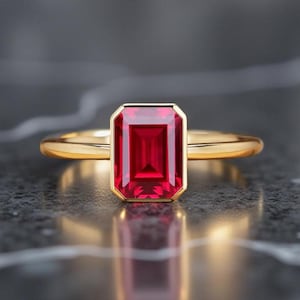 Può includere: Un anello d'oro con una grande gemma rettangolare di colore rosso intenso. La gemma è incastonata in una lunetta d'oro e la fascia è in oro lucido semplice. L'anello è esposto su una superficie scura e riflettente.