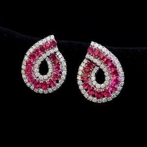 May include: A pair of teardrop-shaped earrings. Each earring features a cluster of small, round, bright pink gemstones surrounded by a border of smaller, clear gemstones. The earrings are set against a black background.