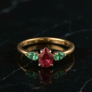 May include: A gold ring featuring a pear-shaped, deep red gemstone flanked by two green, teardrop-shaped stones. The ring is set against a dark background, highlighting the vibrant colors of the gemstones. A beautiful piece of jewelry.
