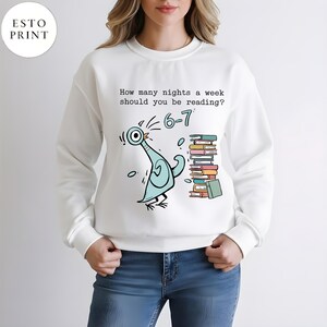 K&ouml;nnte beinhalten: Wei&szlig;es Sweatshirt mit einer Cartoon-Tauben-Illustration und dem Text "How many nights a week should you be reading? 6-7". Die Taube ist hellblau mit schwarzen Akzenten, und ein Stapel B&uuml;cher befindet sich rechts.