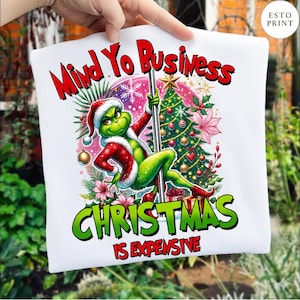May include: White t-shirt with a Grinch graphic in a Santa hat and red suit, pole dancing in front of a Christmas tree. The text reads "Mind Yo Business" and "Christmas Is Expensive" in red and green.