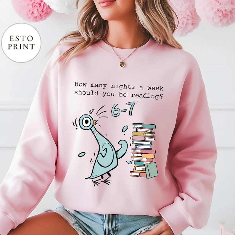 K&ouml;nnte beinhalten: Hellrosa Sweatshirt mit einer Cartoon-Tauben-Illustration und dem Text "How many nights a week should you be reading? 6-7". Die Taube ist hellblau und die B&uuml;cher sind verschiedenfarbig.