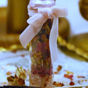 May include: A clear glass bottle filled with dried flowers and salts, tied with a light pink velvet ribbon. The bottle has a light pink lid. The background includes additional dried flowers and vanilla beans.