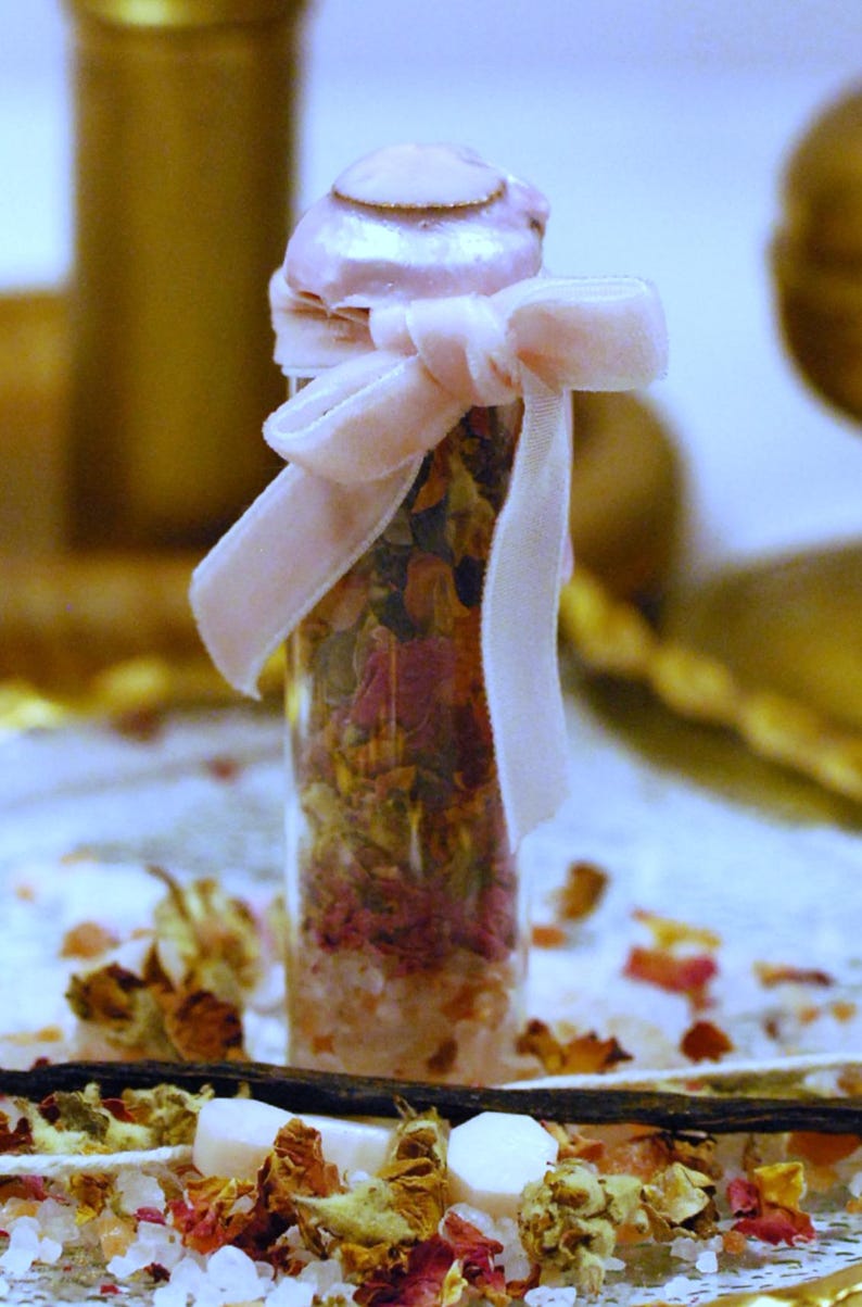May include: A clear glass bottle filled with dried flowers and salts, tied with a light pink velvet ribbon. The bottle has a light pink lid. The background includes additional dried flowers and vanilla beans.