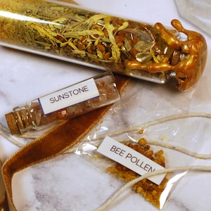 May include: A collection of apothecary items, including a glass vial filled with golden glitter and dried herbs, a small bottle labeled "SUNSTONE", and a packet of "BEE POLLEN". The items are arranged on a marble surface.