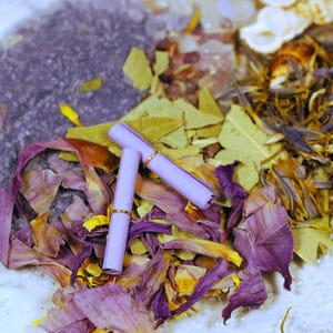 May include: An overhead shot of a collection of dried botanicals and two rolled-up scrolls. The botanicals include purple and yellow flower petals, green leaves, and a dark purple granular substance. The scrolls are light purple with gold rings.