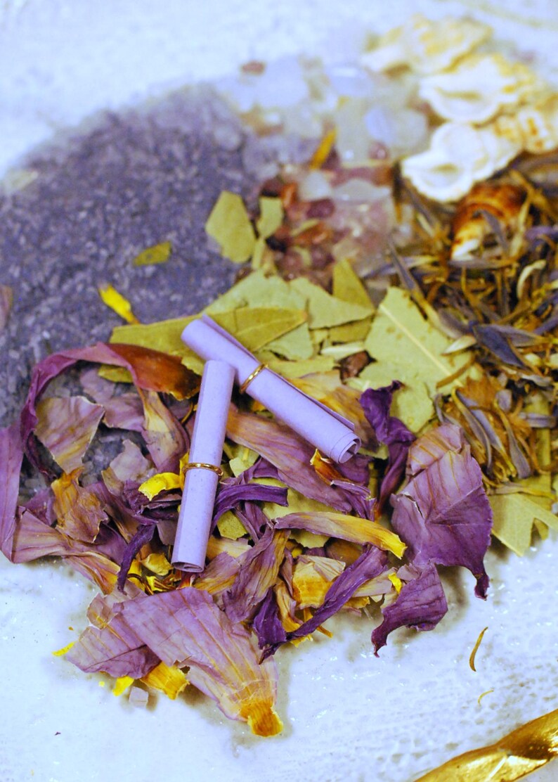 May include: An overhead shot of a collection of dried botanicals and two rolled-up scrolls. The botanicals include purple and yellow flower petals, green leaves, and a dark purple granular substance. The scrolls are light purple with gold rings.