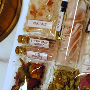 May include: A collection of apothecary items including pink salt, strawberry quartz, rose quartz, rose petals, and apple blossoms. A glass bottle contains pink ribbon. Small bottles of ritual oil are also present. The items are labeled with white labels.