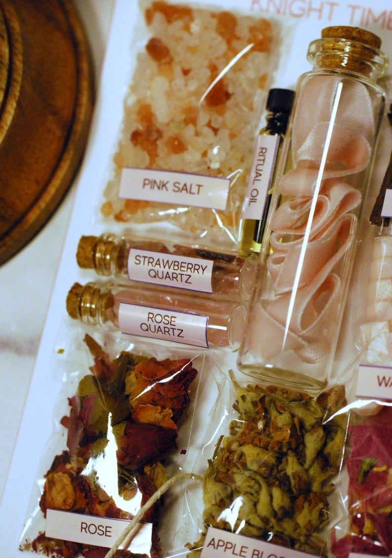 May include: A collection of apothecary items including pink salt, strawberry quartz, rose quartz, rose petals, and apple blossoms. A glass bottle contains pink ribbon. Small bottles of ritual oil are also present. The items are labeled with white labels.