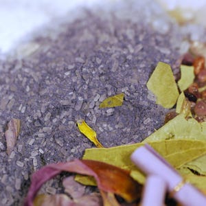 May include: Close-up of a mixture of ingredients, including purple granules, white crystals, and dried leaves. The composition features a variety of textures and colors, with a focus on natural elements. The image suggests a product related to wellness or aromatherapy.