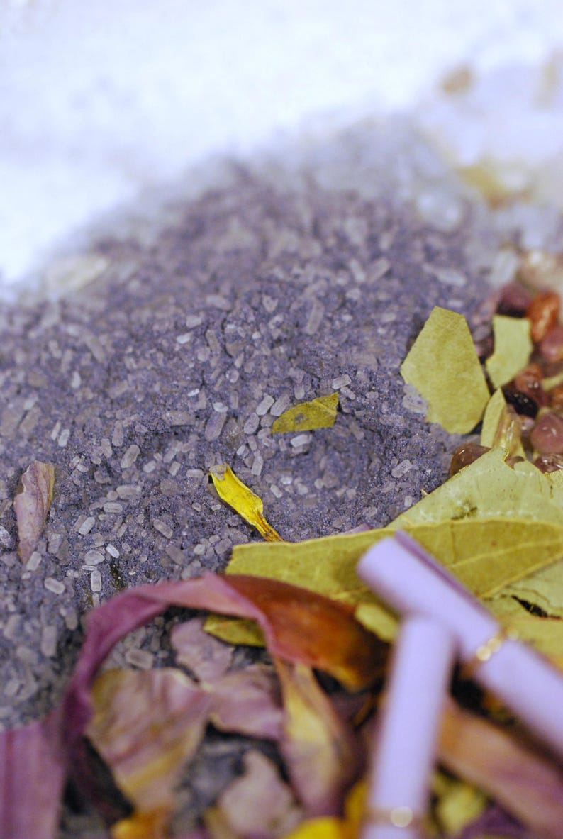 May include: Close-up of a mixture of ingredients, including purple granules, white crystals, and dried leaves. The composition features a variety of textures and colors, with a focus on natural elements. The image suggests a product related to wellness or aromatherapy.