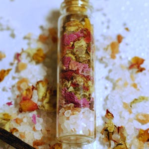 May include: A clear glass bottle filled with white crystals and dried rose petals in shades of pink, red, and yellow. The bottle is sealed with a cork stopper. The background features scattered crystals and petals.
