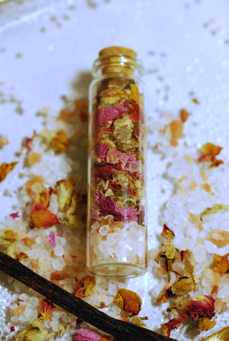 May include: A clear glass bottle filled with white crystals and dried rose petals in shades of pink, red, and yellow. The bottle is sealed with a cork stopper. The background features scattered crystals and petals.