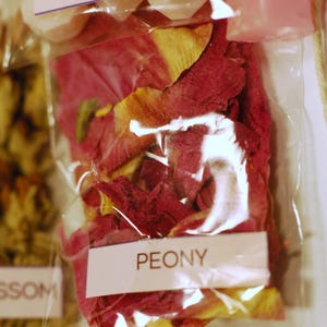 May include: Close-up of clear plastic bags containing wax melts and dried flower petals. One bag is labeled "PEONY" and contains red and yellow petals. Another bag is labeled "WAX MELTS". The image suggests a product for aromatherapy or home fragrance.