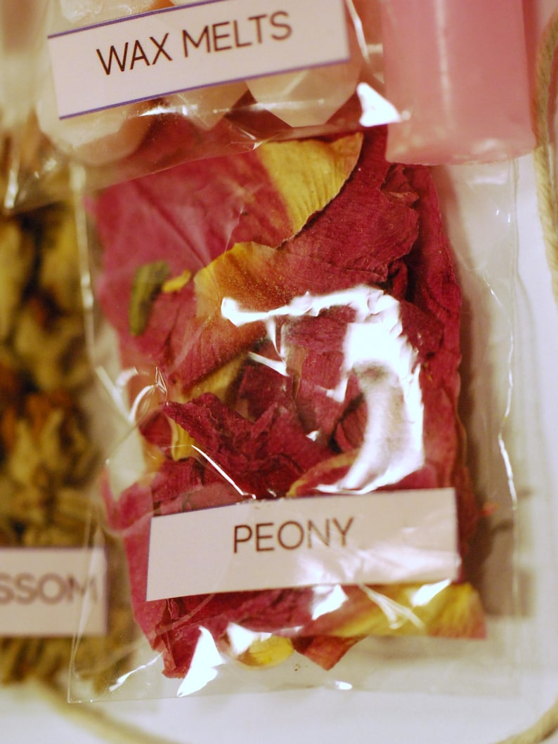 May include: Close-up of clear plastic bags containing wax melts and dried flower petals. One bag is labeled "PEONY" and contains red and yellow petals. Another bag is labeled "WAX MELTS". The image suggests a product for aromatherapy or home fragrance.