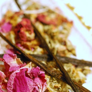 May include: Close-up of a collection of natural ingredients, including pink rose petals, dried flowers, and dark brown vanilla beans. The arrangement is artfully composed, suggesting a focus on aromatherapy or natural beauty products.