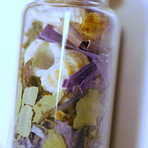 May include: A small, clear glass bottle filled with dried herbs and botanicals. The contents include green, purple, and tan-colored leaves and petals, with a cork stopper. The bottle is likely used for aromatherapy or decorative purposes.