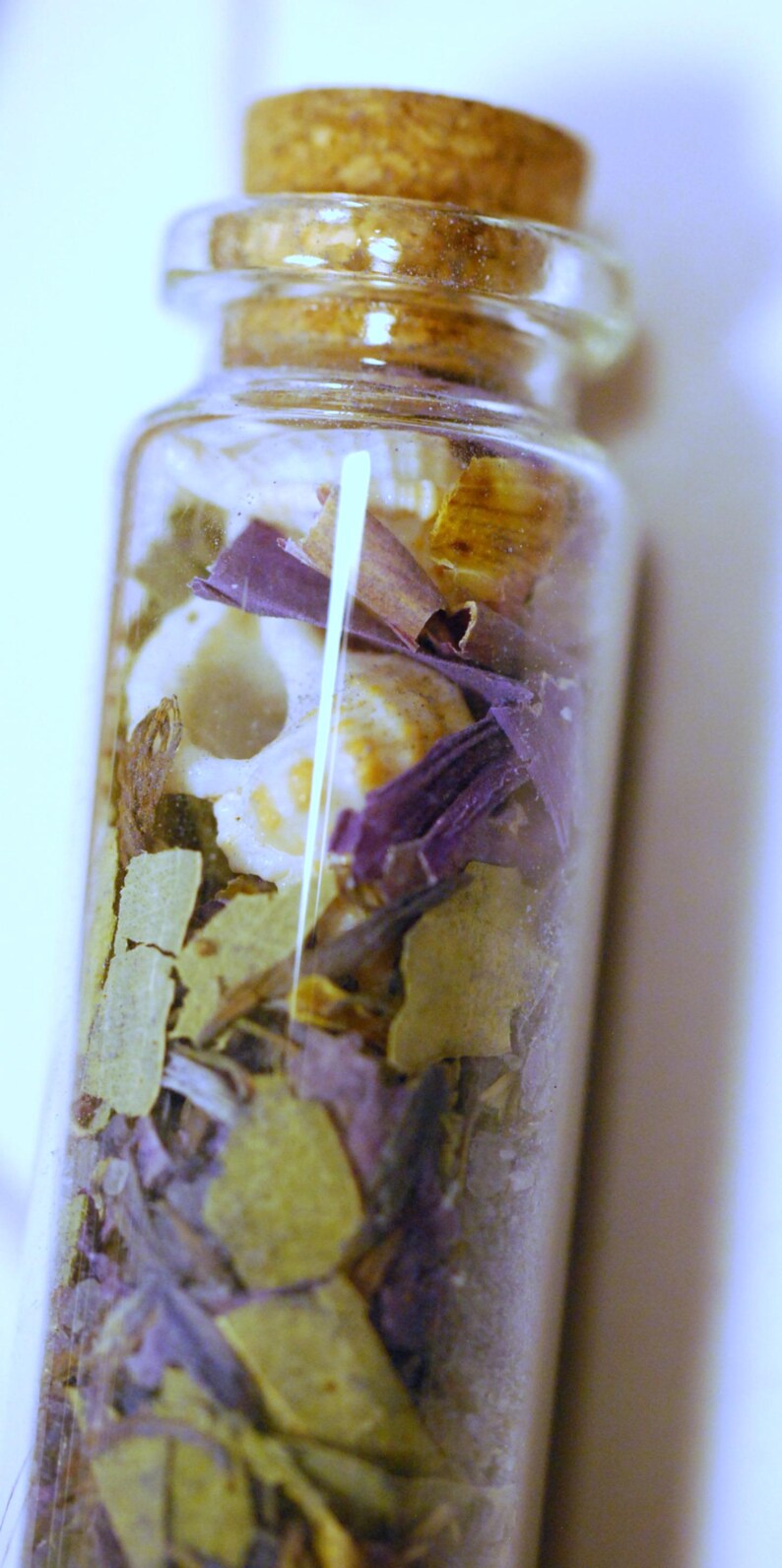 May include: A small, clear glass bottle filled with dried herbs and botanicals. The contents include green, purple, and tan-colored leaves and petals, with a cork stopper. The bottle is likely used for aromatherapy or decorative purposes.