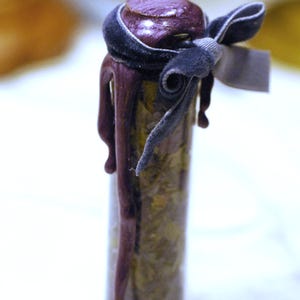 May include: A small glass vial filled with dried herbs, sealed with a dark purple stopper. The stopper is adorned with a dark gray velvet ribbon tied in a bow, and a matching tag. The vial is decorated with dripping purple wax.